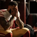 George Kittle sitting on locker room bench with face in hands and golden lighting showing disappointment