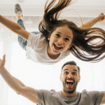 Girl suspended midair in acrobatic pose by long hair with a mischievous grin while father cheers in bright airy room.