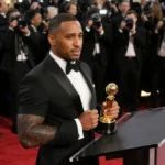 Glen Powell accepts Golden Globe Award at podium with bruised arms and flashing cameras behind