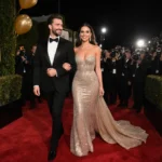 Glen Powell and Michelle Randolph walking hand-in-hand down the red carpet with gold balloons and camera flashes behind