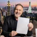 Graham Norton sits with a sly grin holding a blank paper among champagne flutes and wedding invitations in a living room