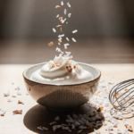 Bowl of creamy Greek yogurt being sprinkled with coconut flakes with soft natural light from above on a minimalist Japanese w