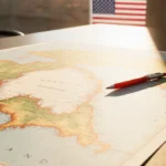 Hand traces Greenland map outline with red pen while American flag fades in background under warm golden light