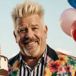 Guy Fieri poses with spiky blonde hair and bold striped shirt beside a small birthday cake.