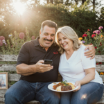 Guy Fieri smiles while checking his phone with Lori beside him holding a plate of food in their backyard.