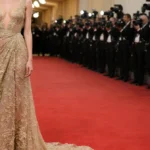 Hailey Bieber walking red carpet with Armani Privé lace gown and golden glow against blurred cameras