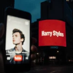 Harry Styles fan watching new music video on phone with billboard showing album artwork above city street