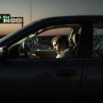 Sedan parked with a lifeless body on passenger seat and shattered glass amid highway dawn