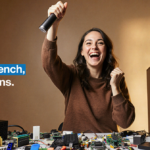 Excited person holding up a Dell desktop with a big smile against a cluttered desk full of accessories