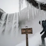 Hiker carefully steps around large icicle on Mt Baldy trail with fog swirling around legs and caution sign showing icy condit