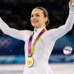 Ilia Malinin celebrates Olympic gold victory with arms raised and medal gleaming against blurred ice rink