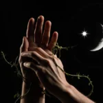 Two pairs of hands intertwine with fingers wrapped together under dim lighting with moon and planets visible behind