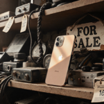 iPhone 16 rests on a cluttered second‑hand shelf with warm light on its screen and a crooked