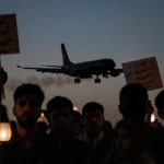 Protesters holding signs confront security forces at airport with black military plane taking off in background