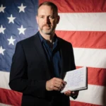 Jack Smith standing confidently with small notebook showing deposition notes against a patriotic flag backdrop determined