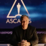 James Cameron sits confidently at desk with Academy Awards logo glowing behind him and trophy case beside him
