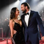 Jason Momoa and Adria Arjona posing together on red carpet with her black gown and his navy tuxedo glowing under spotlights