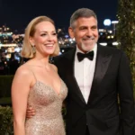 Jean Smart and George Clooney smiling on the red carpet with golden decorations and city lights behind them