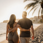Jennifer Aniston and Jim Curtis walking hand‑in‑hand along Mediterranean beach at sunset with crystal‑clear water palm trees