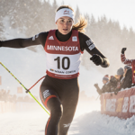Jessie Diggins mid-stride with arms outstretched and determined expression against Olympic winter skiing spectators cheer