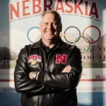 Jim Hartung stands with crossed arms and warm smile wearing Husker jacket with Nebraska gymnastics backdrop and faded Olympic