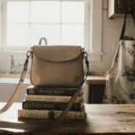 Joanna Gaines style crossbody bag rests on vintage cookbooks with distressed apron hanging nearby and soft window light filli