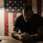 Joe Rogan sits with clasped hands at worn desk with vintage microphone and American flag in dimly lit studio