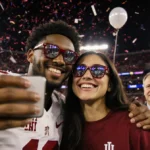 Jordon Hudson and Alix Earle smile while taking a selfie with phones and confetti in a neon-lit college championship stadium.