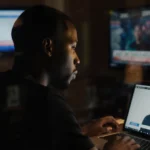 Karamo Brown staring at laptop screen showing a video message with soft warm lighting and blurred TV screens in background
