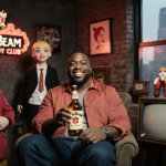 Kenan Thompson smiles holding a Jim Beam bottle with nostalgic Nickelodeon props and a city skyline in the background