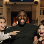 Daughters laughing and pointing at Kenan Thompson while he watches TV in a cozy living room with soft golden light