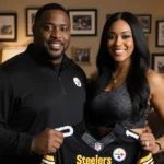 Kiya Tomlin stands with Mike Tomlin holding Steelers jersey with family photos and sports memorabilia on wall behind them