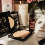 Laura Geller compact powder sits on organized beauty shelf with skincare products and makeup arranged nearby