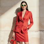 Lauren Sanchez Bezos stands confidently with a red skirt suit and gradient aviator sunglasses against a neutral backdrop.