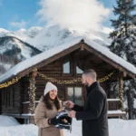 Leslie Davis smiles while holding a snowmobile helmet as her fiancé hands her keys with a cozy cabin and snowy mountains behi