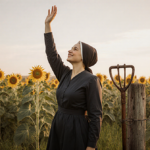Lizzie Ens standing in a green field with sunflowers extending her arm upward and smiling embodying freedom and empowerment.