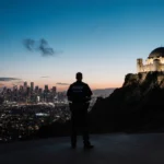 Lone figure stands under Griffith Observatory at dusk with LA skyline glowing and police patrol car visible in distance