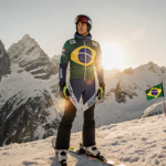 Lucas Pinheiro Braathen stands with skis at his feet, wearing a suit and looking determined as sunset lights mountain with fl