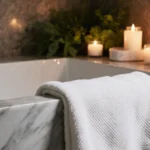 White luxury bath towel drapes across marble tub with candlelight and greenery creating spa atmosphere