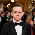 Macaulay Culkin walking red carpet in black tuxedo with golden lights and champagne glasses behind him