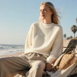 Woman relaxes on weathered bench wearing ivory sweater with palm trees and ocean waves behind