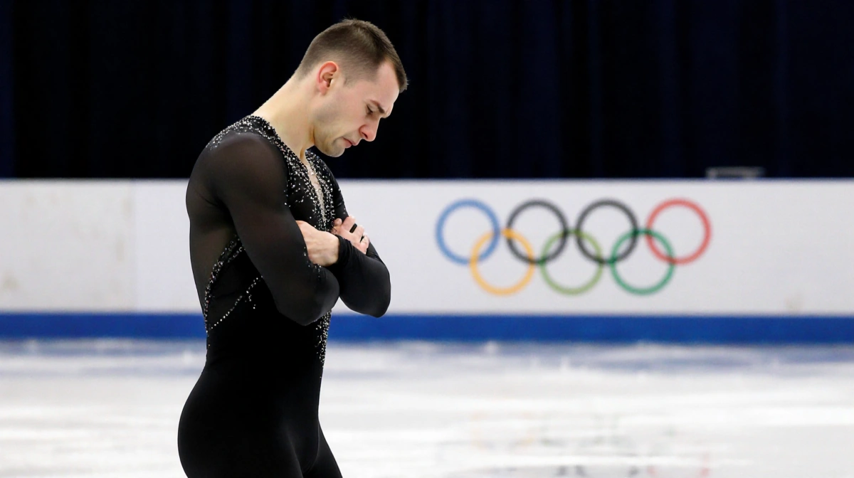 Ilia Malinin stands with downcast eyes and arms crossed tight showing Olympic rings behind him