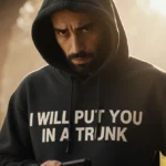 Man wearing hoodie with threatening message looking concerned while holding phone and standing with blurred background