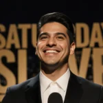Marcello Hernández standing at microphone with Saturday Night Live logo behind him and golden glow illuminating face