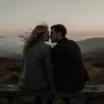 Margot Robbie and Jacob Elordi sit together on a worn stone bench at dusk with misty moorland behind them sharing an intimate