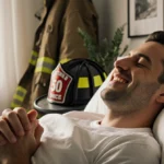 Matthew sitting up in bed with closed eyes and sunlight streaming through window showing firefighter helmet nearby
