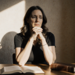 Melissa Gilbert sits at a desk with hands clasped natural light illuminating her face and an open leather‑bound journal besid