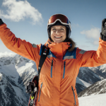 Meredith Hayden smiles with arms outstretched and fist raised at a snowy mountain summit with bright orange jacket and skis t