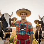 Mexican farmer standing with two black-and-white cows wearing cowbell necklaces and wildflowers in a green meadow.