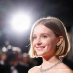 Millie Bobby Brown smiling on red carpet with short bob and diamond necklace under spotlight